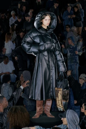 Fashionable woman in puffy black coat stands out in a dark, crowded scene, holding a tote bag, captured in a full-length shot.