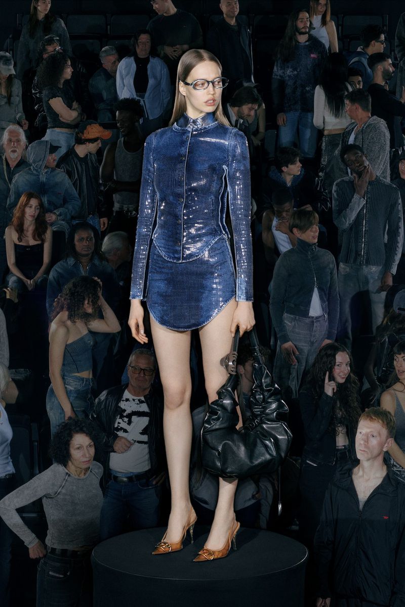 Fashion show with a female model wearing a blue sequined dress and glasses on a round stage, with a large audience in the background.