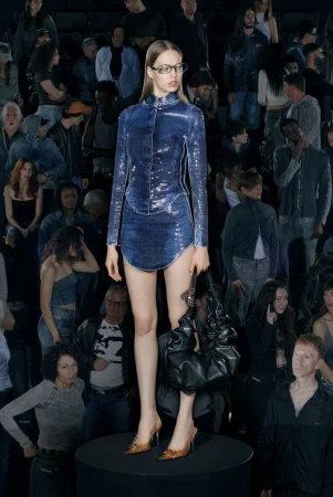 Fashion show with a female model wearing a blue sequined dress and glasses on a round stage, with a large audience in the background.