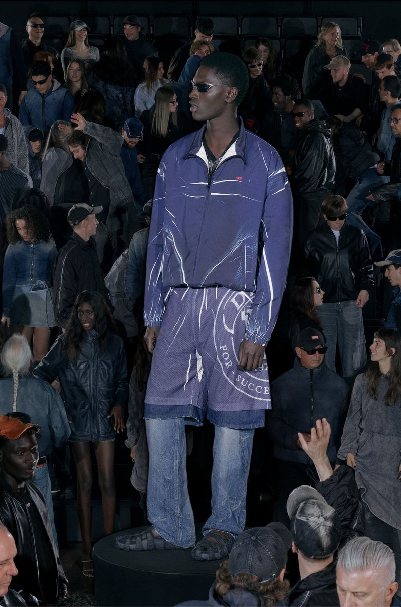 Fashion show runway featuring a model in a blue jacket and shorts, surrounded by a crowd, in a dimly lit, indoor setting.