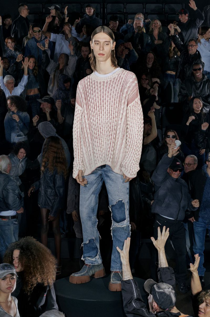 Fashion model stands on a black platform wearing a cable-knit sweater and distressed denim jeans, surrounded by a blurred crowd.
