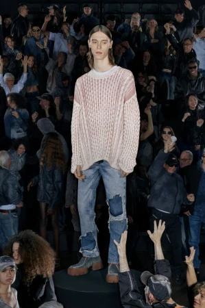 Fashion model stands on a black platform wearing a cable-knit sweater and distressed denim jeans, surrounded by a blurred crowd.