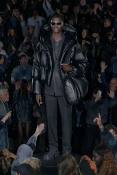 Model wearing a black puffer jacket and sunglasses on a runway surrounded by a seated crowd for Diesel Resort 26