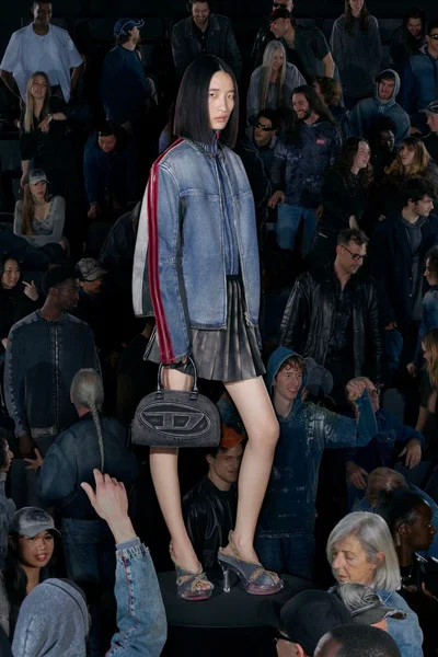 Model wearing a denim jacket and pleated skirt holding a handbag on runway for Diesel Resort 26