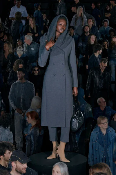 Model standing on a circular runway wearing a grey hooded coat and trousers surrounded by an audience
