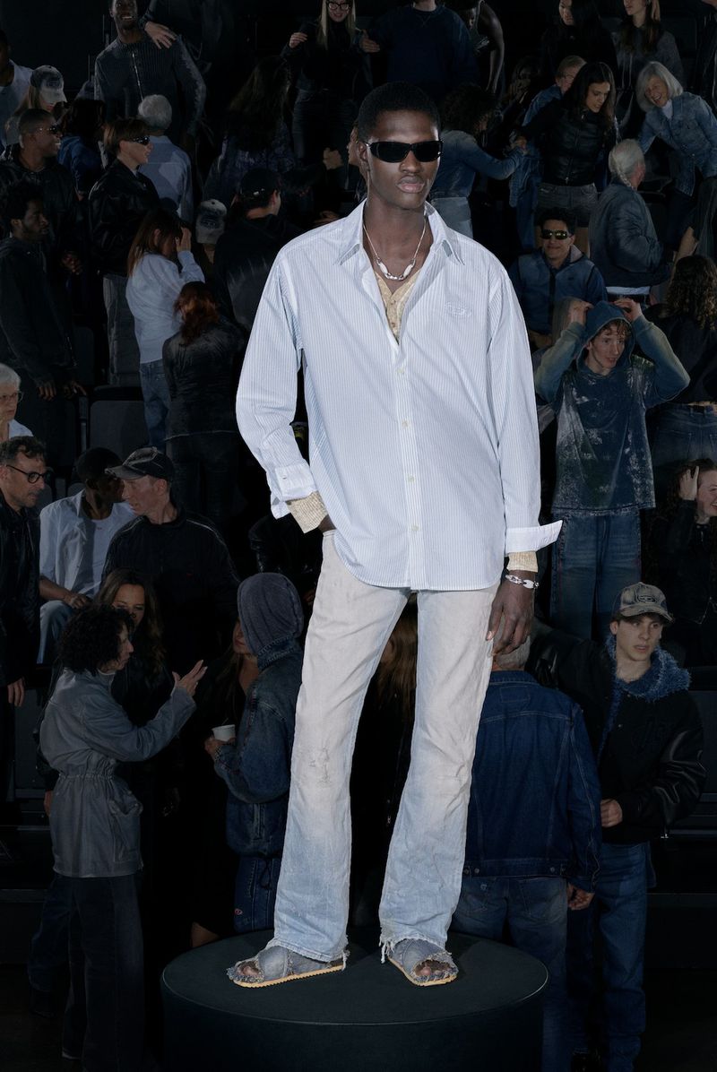 Model wearing a light blue button-down shirt and light jeans on a round pedestal before a seated audience