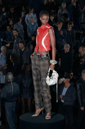 Model standing on a circular podium wearing a red zip vest and patterned trousers holding a white handbag with audience in background