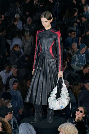 Model wearing a black and red leather jacket and pleated skirt holding a patterned handbag on a runway