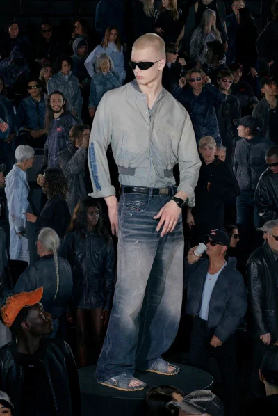 Model standing on a circular podium wearing sunglasses, grey shirt, and wide-leg denim jeans amid a crowd