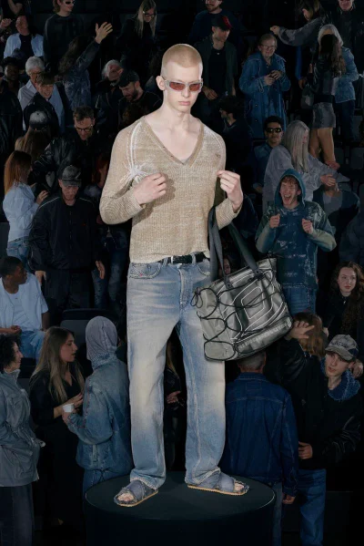 Model standing on a circular podium wearing tinted sunglasses, beige V-neck sweater, light wash jeans, holding a grey tote bag