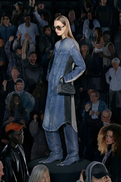 Model standing on a circular podium wearing a long denim coat over matching denim boots, holding a small handbag against a crowd background
