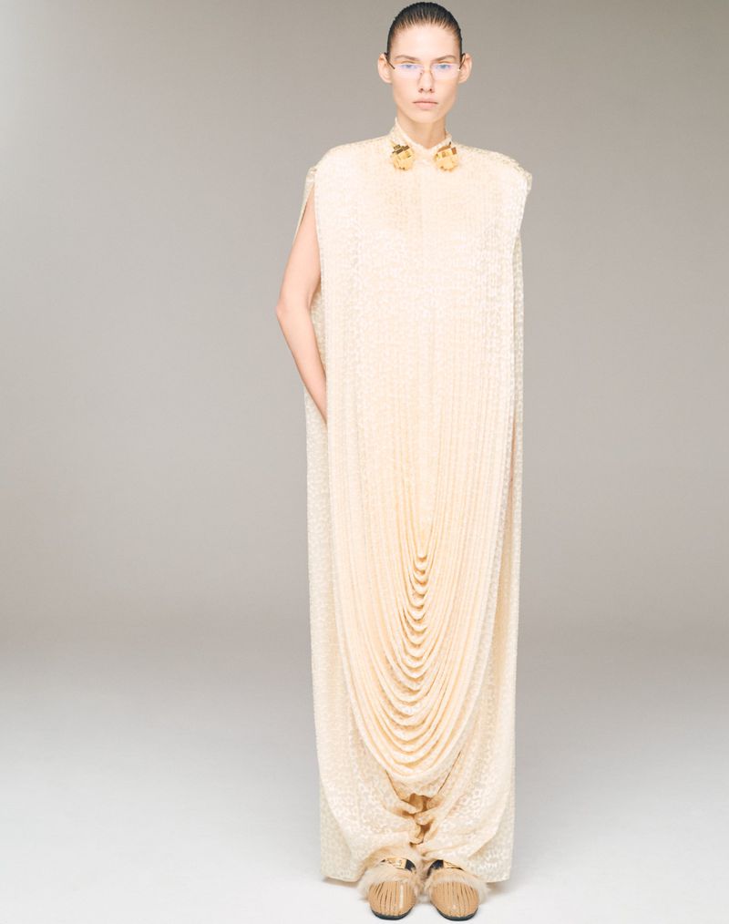 A full studio shot of a slim adult woman modeling a long, light-beige gown with an elaborate draped front and beaded collar and shoulders.