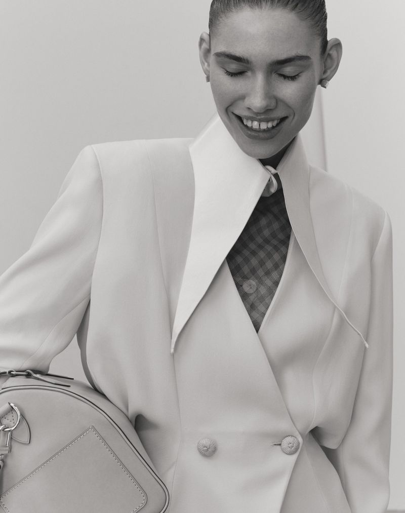 Model smiling while wearing a structured white jacket and holding a handbag against a plain background