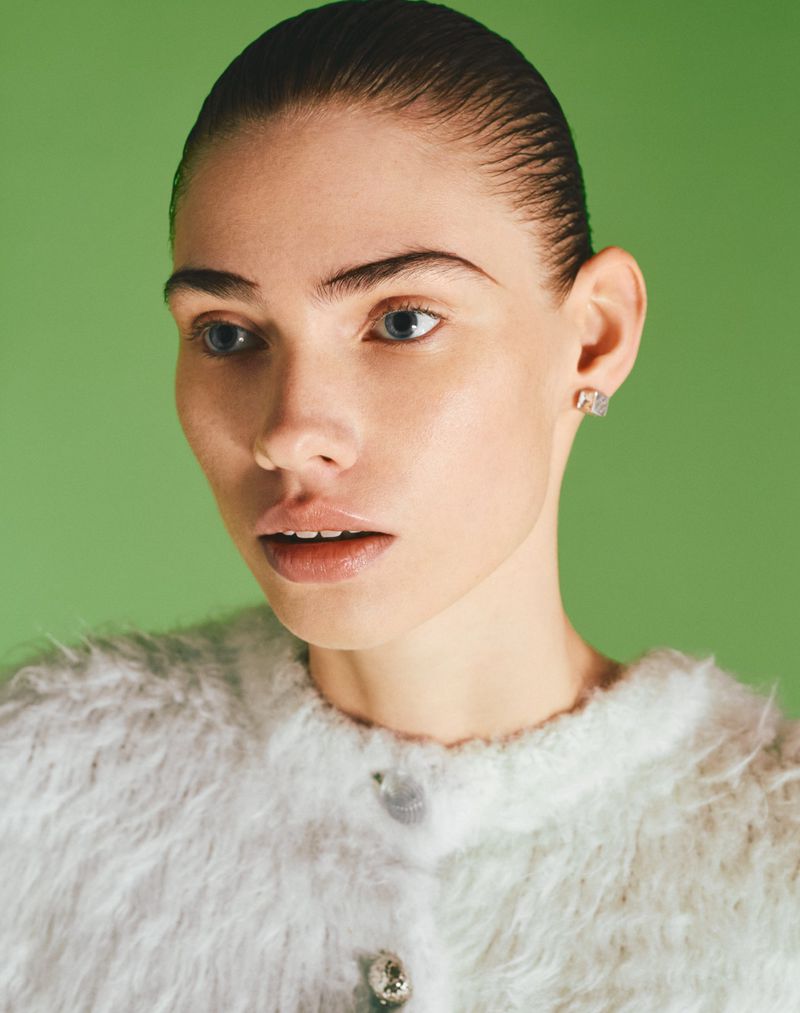 Model with slicked-back dark hair wearing a white textured sweater against a green background