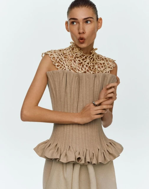 Fashion editorial shot of a young adult woman in a neutral color ribbed corset top and decorative collar against a white backdrop.