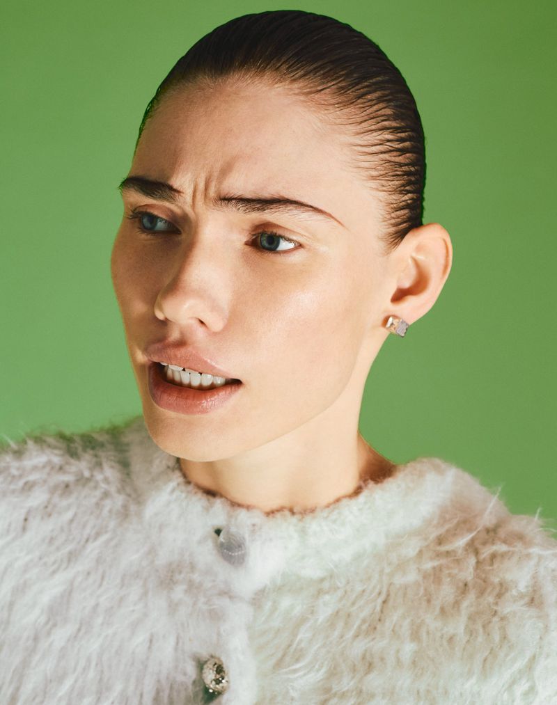 Close-up portrait of a woman with slicked-back hair and wearing a white, fuzzy cardigan against a vibrant green backdrop. Fashion shot.