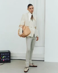 Model Cara Schädel wearing a structured white jacket and grey trousers holding a tan handbag in a studio setting for Vogue Turkey.