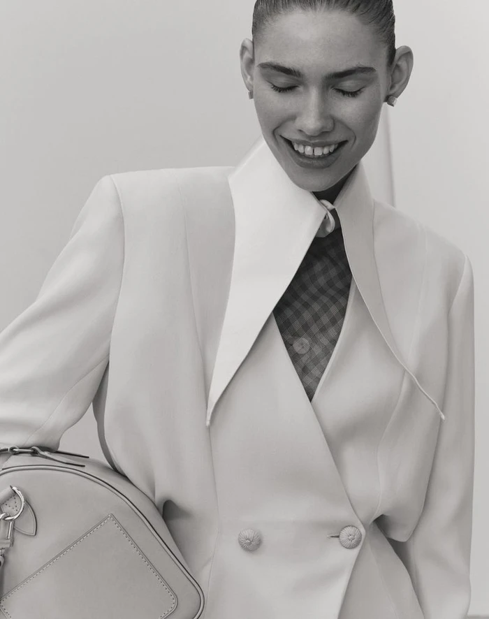 Model smiling while wearing a structured white jacket and holding a handbag against a plain background