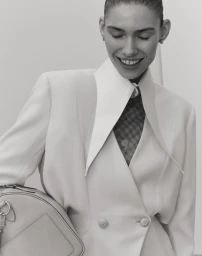 Model smiling while wearing a structured white jacket and holding a handbag against a plain background