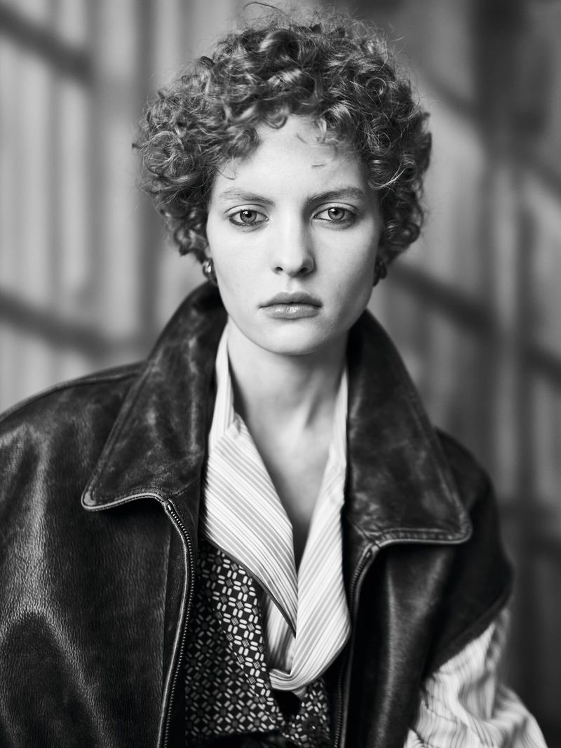 Black and white portrait of an adult female with curly hair, wearing a leather jacket and patterned shirt, against a neutral backdrop.
