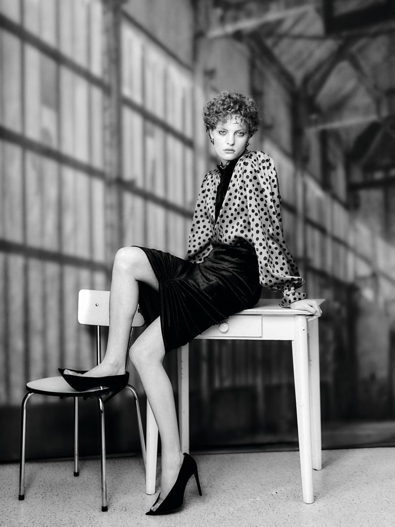 Fashionable woman sits on a white table in a studio wearing black heels and a polka dot blouse in a black and white photo.