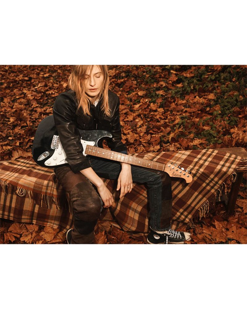 A young woman with a guitar sits on a bench covered with a blanket, outdoors in autumn foliage, wearing jeans, a black jacket, and sneakers.