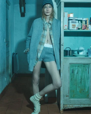 A fashion photo showcases a slender person wearing a knit hat and cardigan in a kitchen interior. Cool lighting highlights a vintage aesthetic.