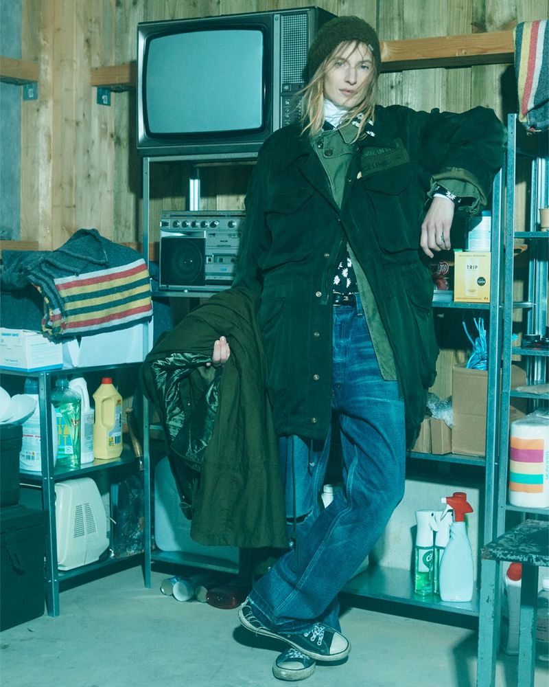 Person in a green jacket and blue jeans stands in a cluttered garage, with shelves, an old television, a boombox, and cleaning supplies.