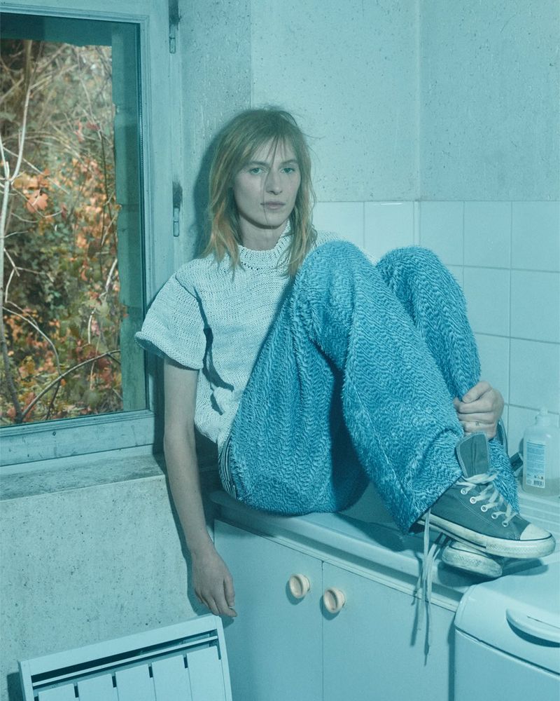 A woman with long hair wearing wide-leg blue textured pants and a matching top sits on the edge of a white tiled counter indoors.
