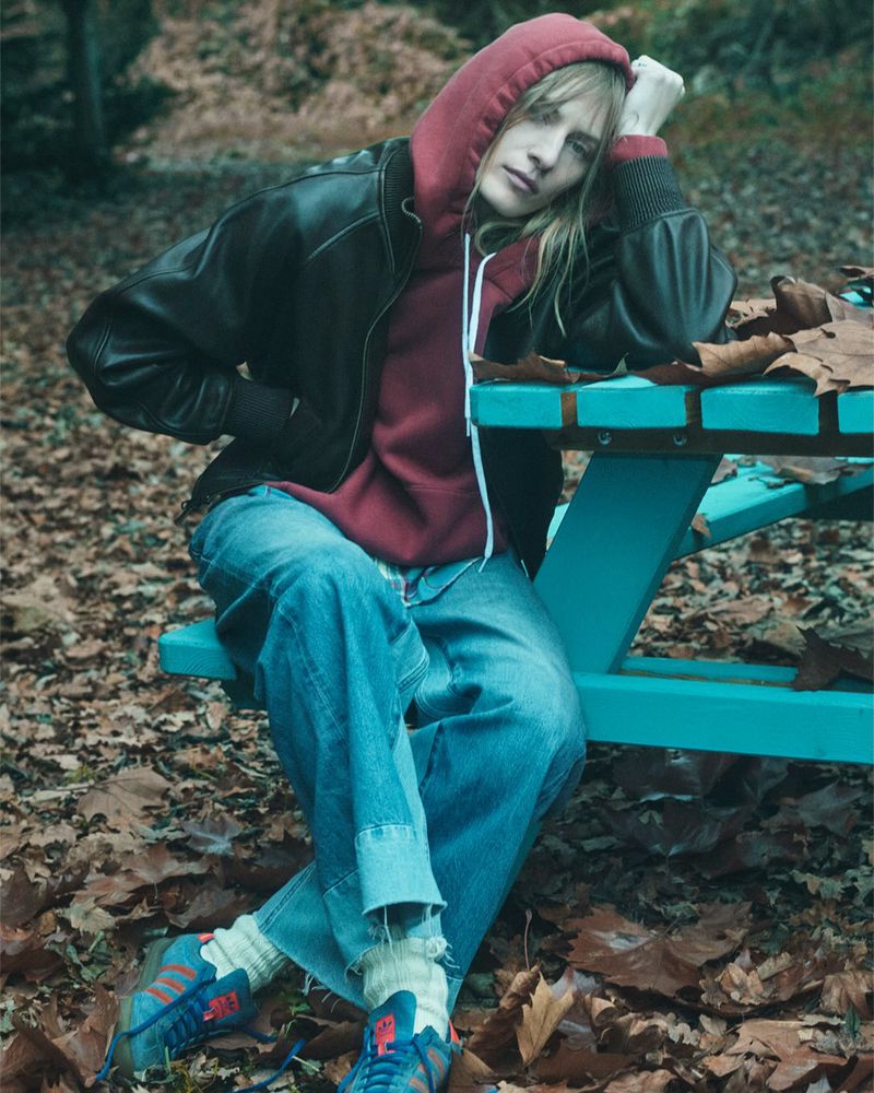 A young adult woman sits on a teal picnic table in an outdoor setting, wearing a hoodie, leather jacket, jeans, and sneakers.