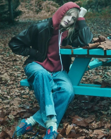 A young adult woman sits on a teal picnic table in an outdoor setting, wearing a hoodie, leather jacket, jeans, and sneakers.