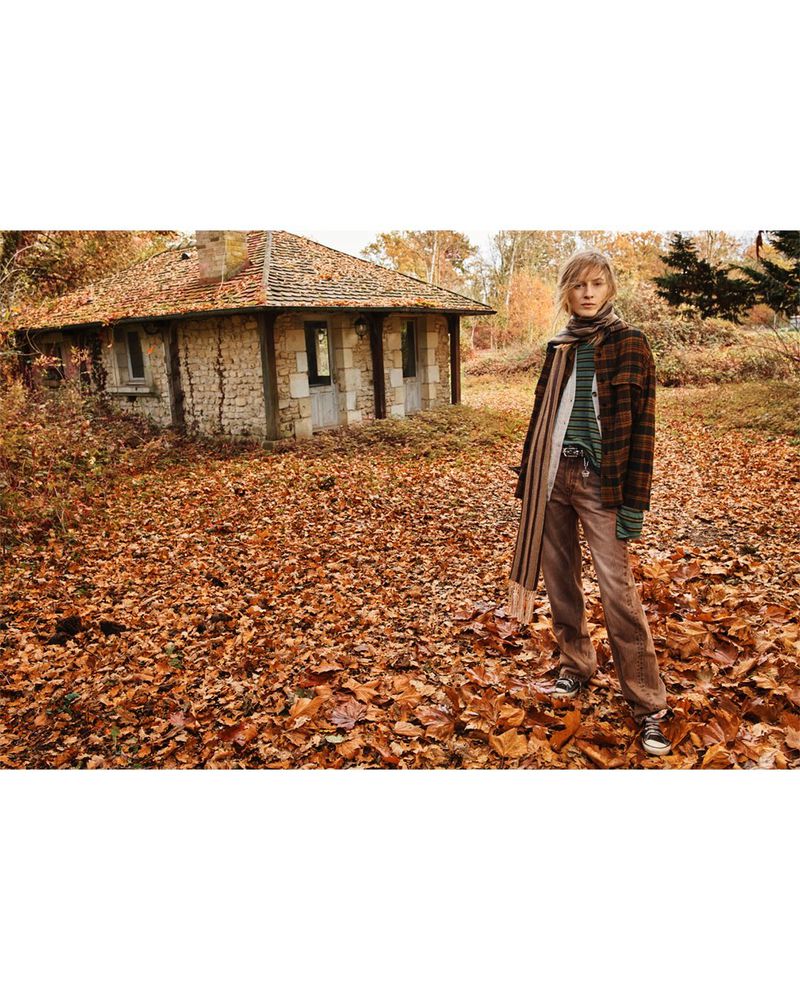Female model poses in fall foliage wearing a patterned jacket and scarf, with a stone building visible in the background in an outdoor setting.