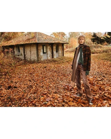 Female model poses in fall foliage wearing a patterned jacket and scarf, with a stone building visible in the background in an outdoor setting.