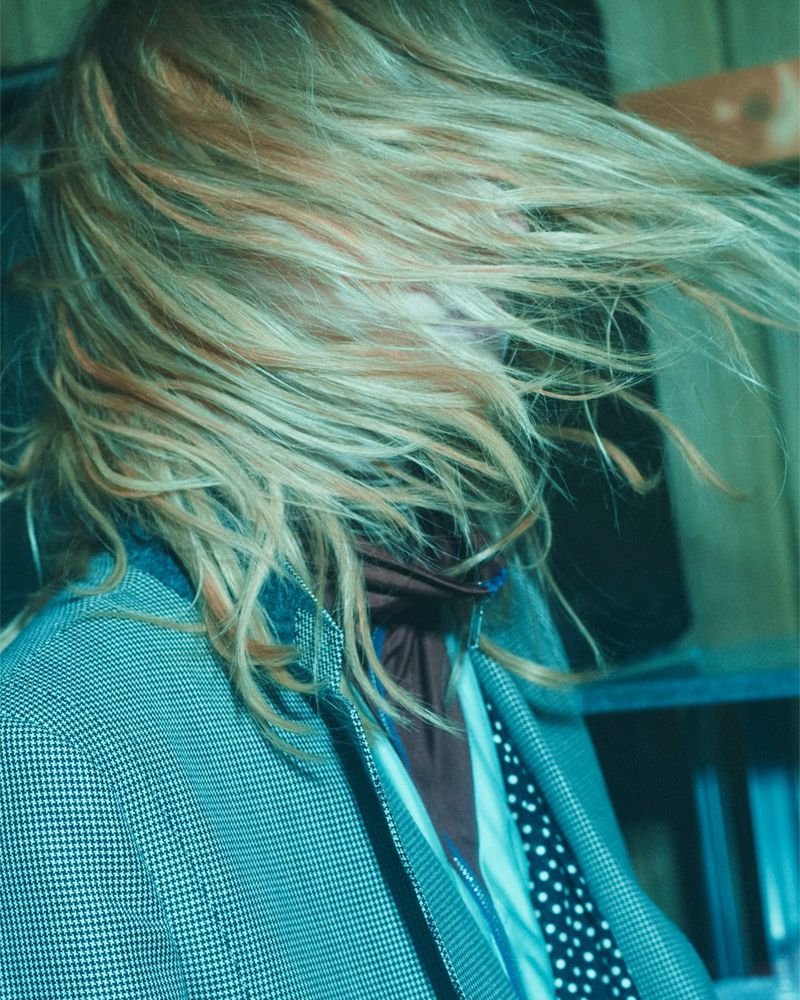 Womans face obscured by blonde hair, wearing a grey jacket and black-and-white polka dot necktie in front of a dark grey shelving unit.