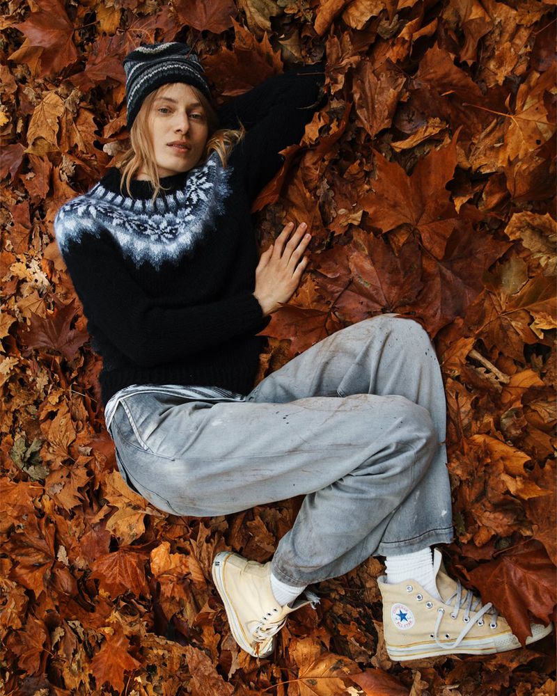 Woman in a patterned sweater and beanie lies in a pile of golden autumn leaves, wearing Converse sneakers, creating a cozy seasonal scene.