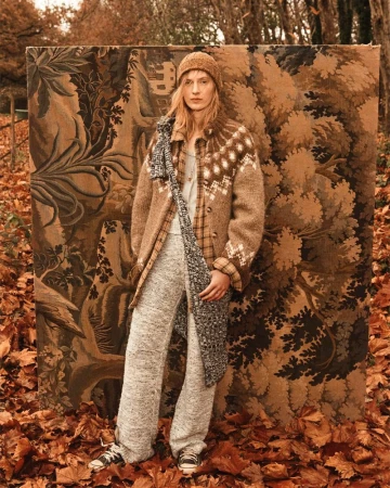 A woman wearing a knit hat, coat, and sweatpants stands in front of a tapestry on a bed of autumn leaves in an outdoor setting.