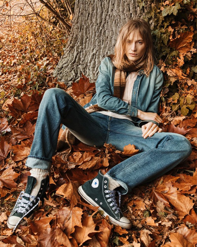 Adult female with long blonde hair wearing a denim jacket and jeans, posing in fallen autumn leaves near a tree in outdoor setting.