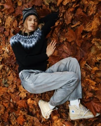 Model Julia Nobis lying amidst autumn leaves wearing a patterned black knit sweater and beanie