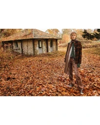 Model Julia Nobis wearing a plaid jacket and scarf stands amidst fallen autumn leaves before a stone house