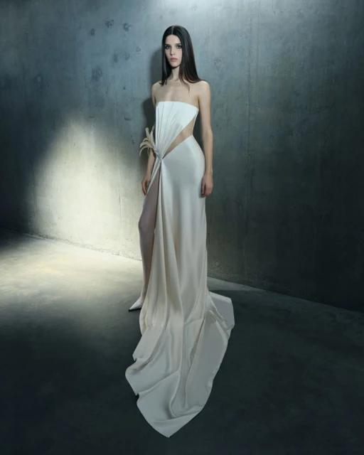 A woman poses in a long strapless white gown with a high slit in a studio setting. The woman is in the center of the frame.