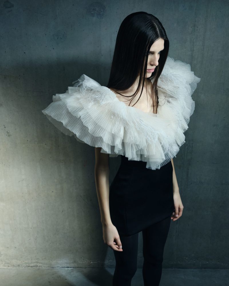 A stylish woman with long dark hair poses in a black dress with a large, layered white ruffled collar against a textured grey wall.