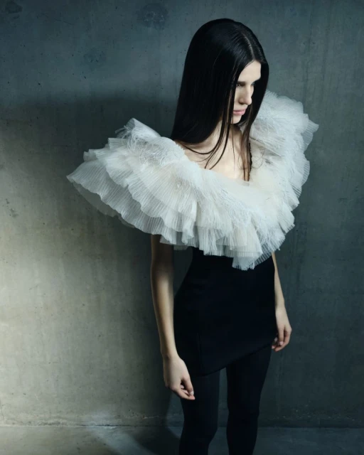 A stylish woman with long dark hair poses in a black dress with a large, layered white ruffled collar against a textured grey wall.
