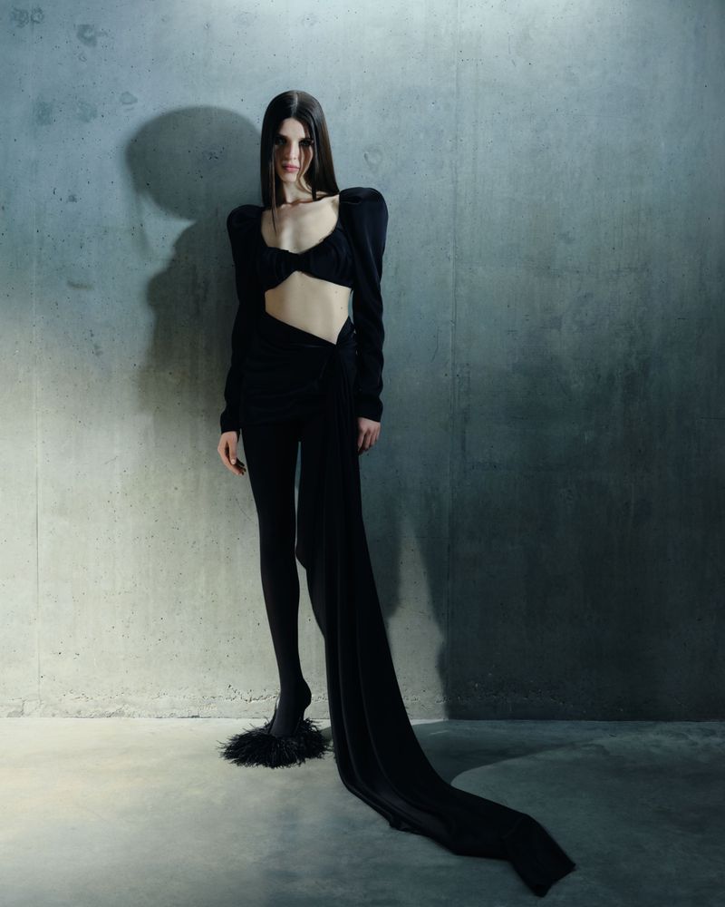 A slender woman in a black two-piece outfit with long sleeves poses in front of a textured grey wall in a fashion forward pose.