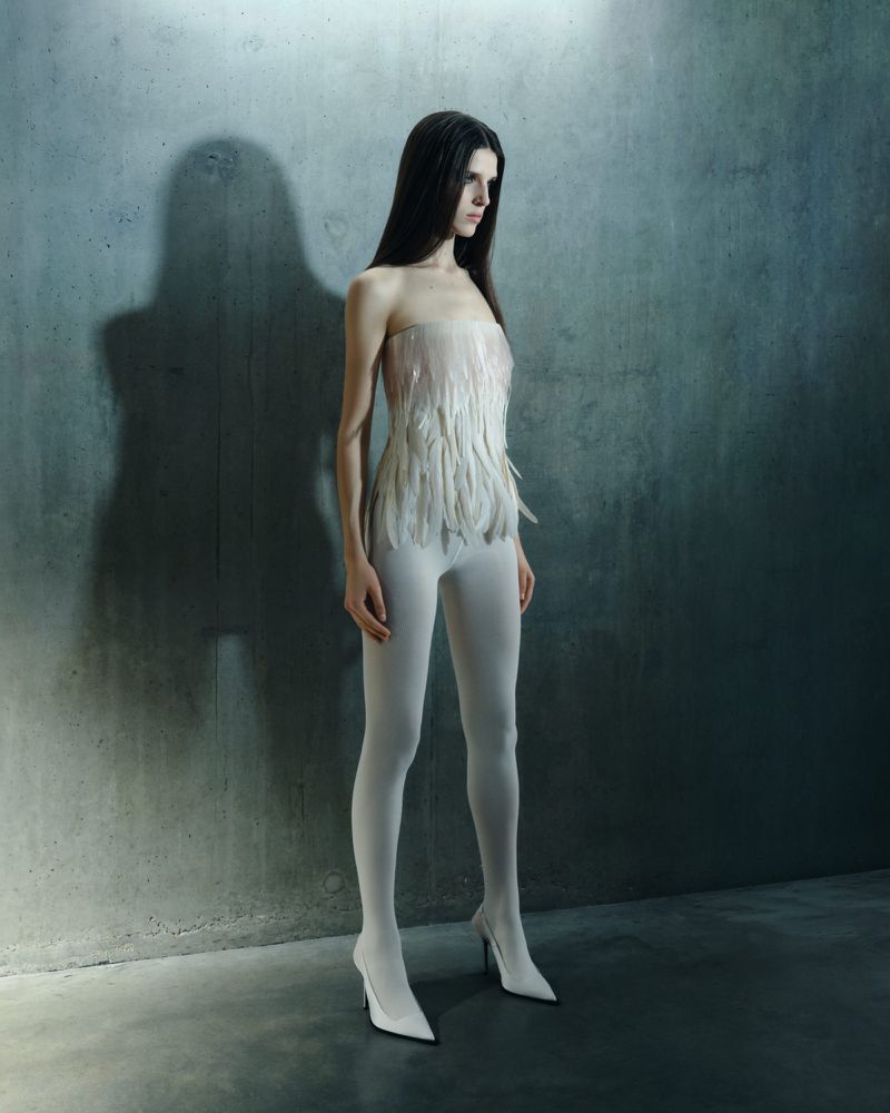 A woman stands against a textured gray wall, wearing a feather top, tights, and heels, illuminated by stark lighting and a dark shadow.