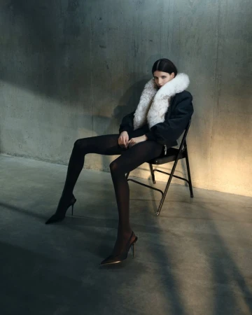 An adult female fashion model sits on a black folding chair in a studio wearing a black jacket, black tights, and heels.
