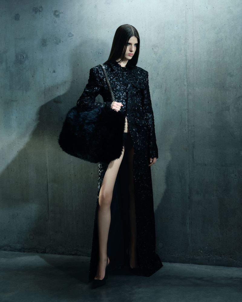 Fashion model poses in a long black sequined coat with a faux fur bag against a gray concrete background in a studio fashion shot.