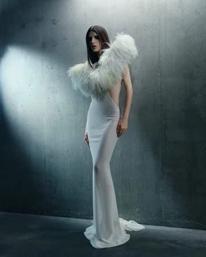 Model wearing a white dress with a feathered collar against a textured concrete wall under cool studio lighting.