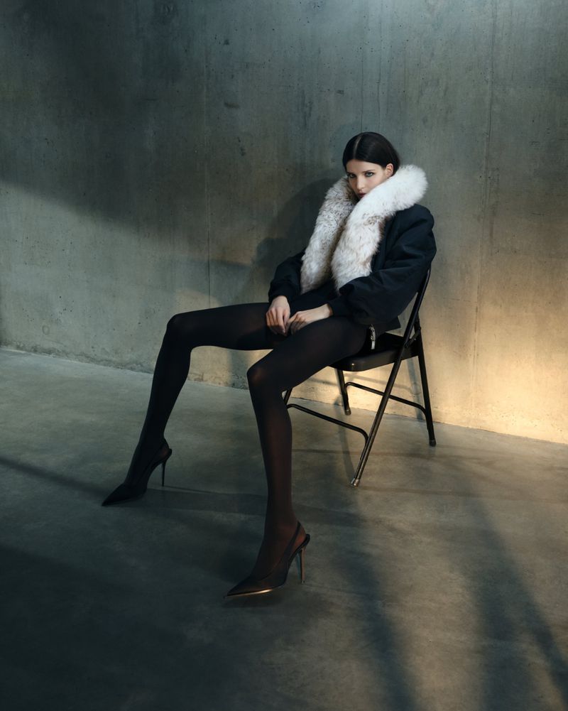 Model seated on a black folding chair against a concrete wall wearing a dark jacket with a white fur collar