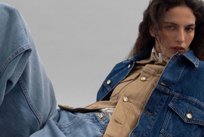 A brunette woman poses in a denim jacket and jeans against a gray background, showcasing a casual yet fashionable look.