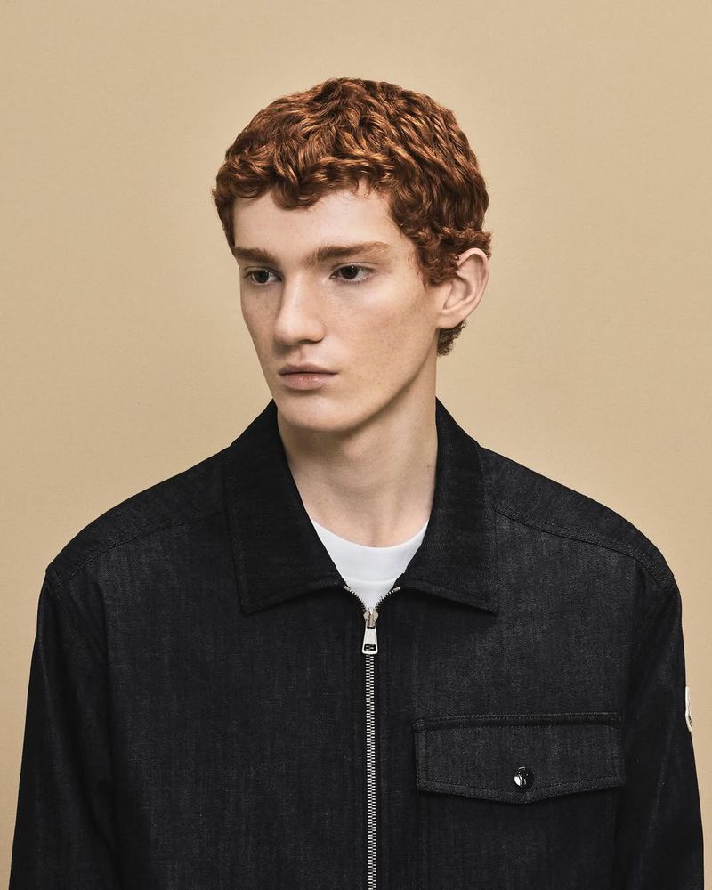 A young adult man with red curly hair poses in a dark denim zip-up shirt against a beige studio backdrop for a fashion portrait.